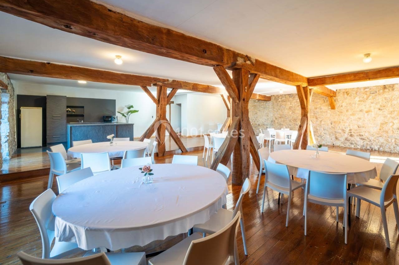 Salle lumineuse avec poutres apparentes, tables rondes dressées de nappes blanches et coin cuisine en arrière-plan.