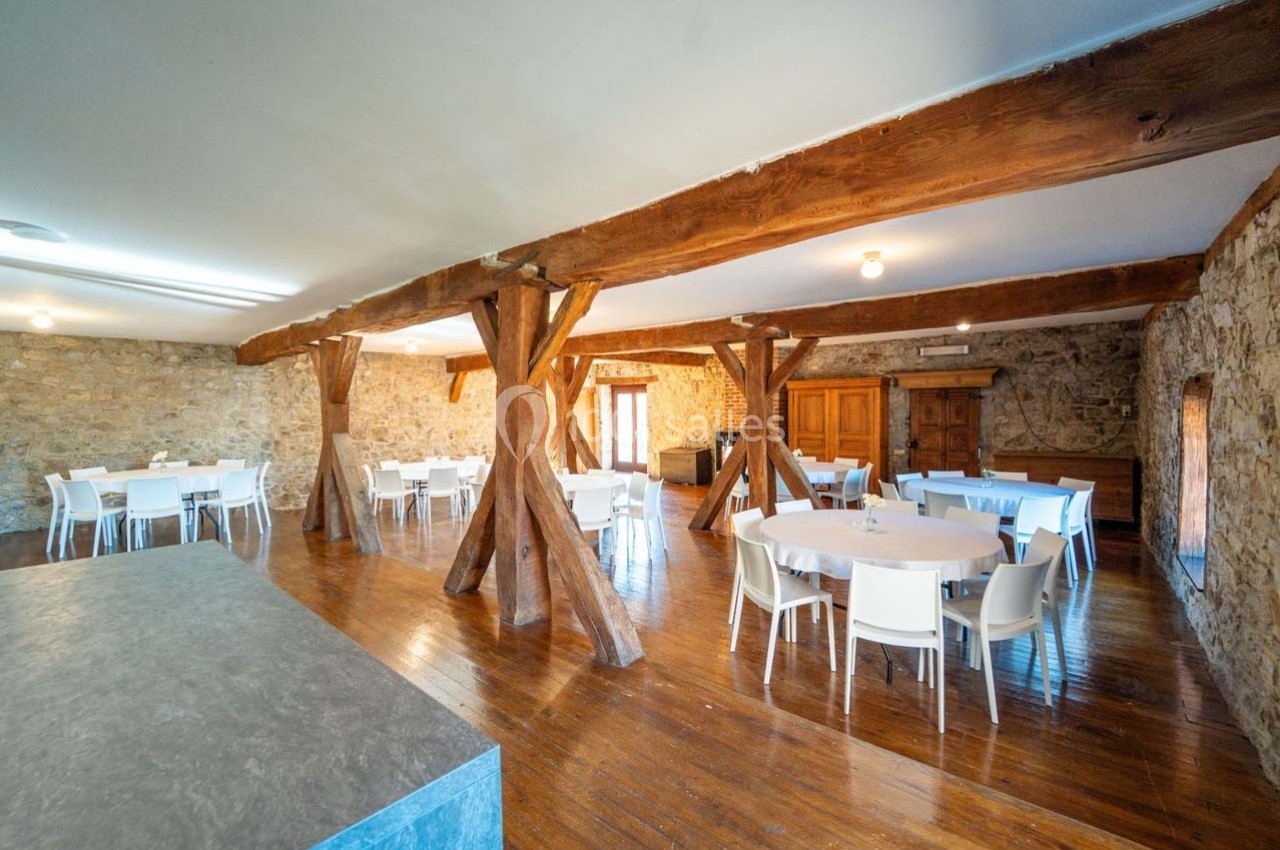 Salle spacieuse avec murs en pierre, poutres en bois apparentes, tables rondes blanches et chaises disposées.