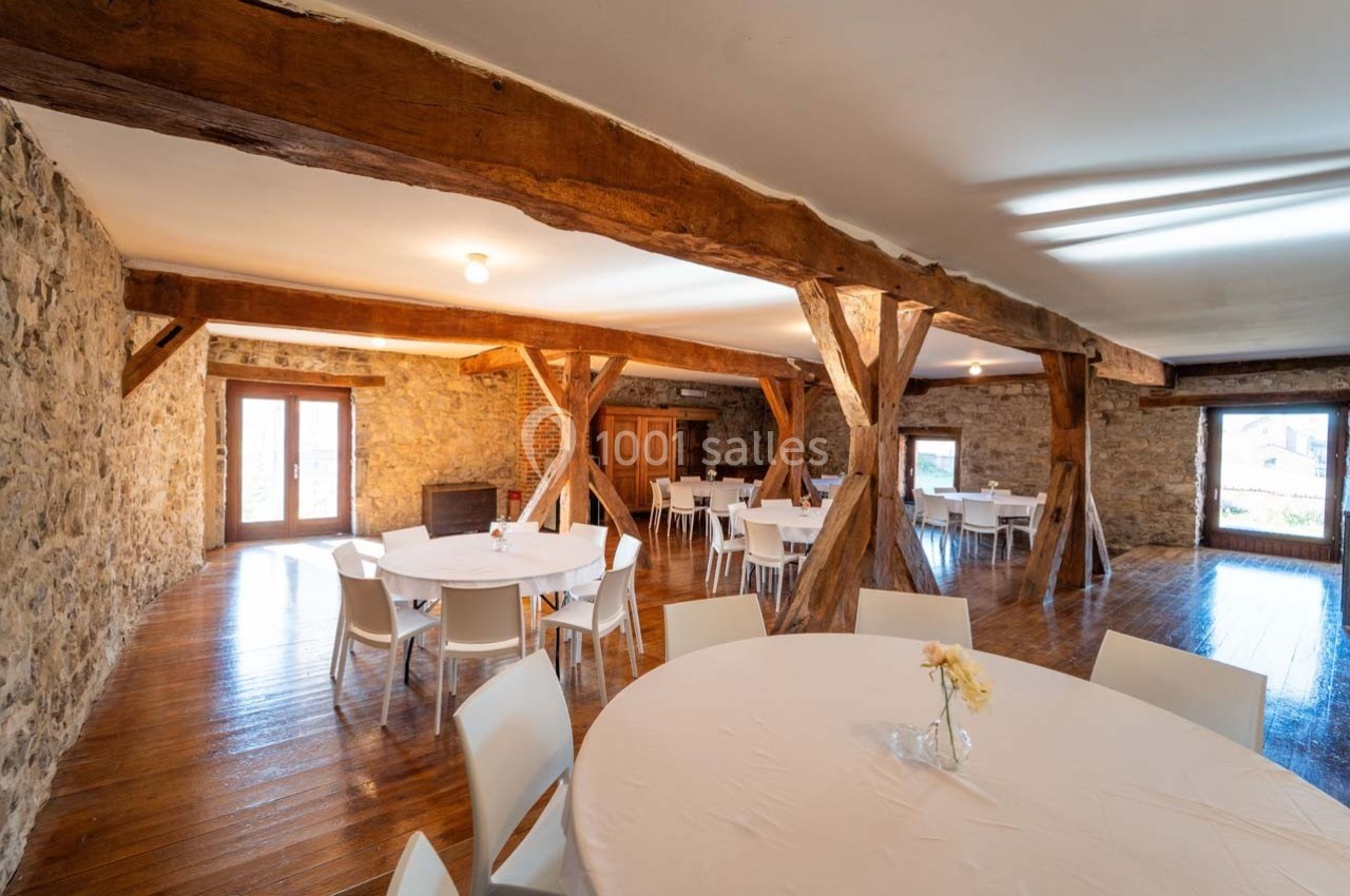 Salle lumineuse avec murs en pierre, poutres apparentes, tables rondes blanches et chaises, vue sur l'extérieur.