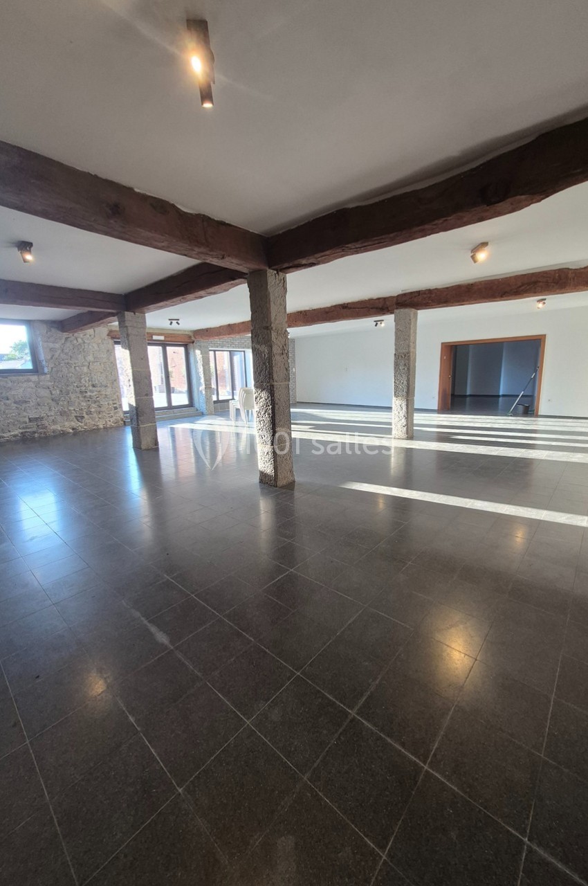 Salle spacieuse avec sol en carrelage sombre, murs en pierre et poutres apparentes, éclairée par des fenêtres.