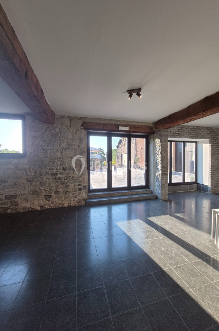 Salle lumineuse avec murs en pierre, poutres apparentes, sol en carrelage sombre et grandes baies vitrées donnant sur l…