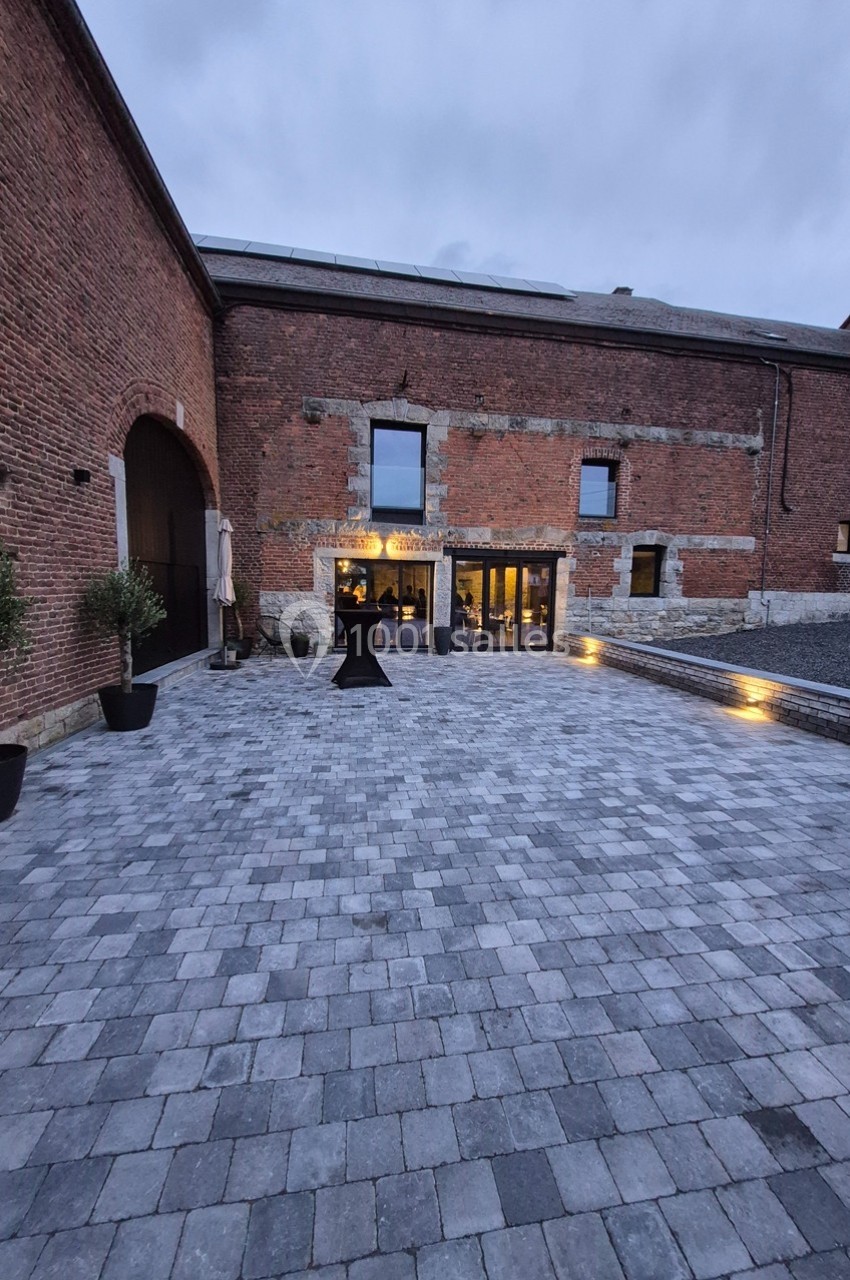 Cour pavée devant un bâtiment en briques avec grandes fenêtres illuminées à la tombée de la nuit.