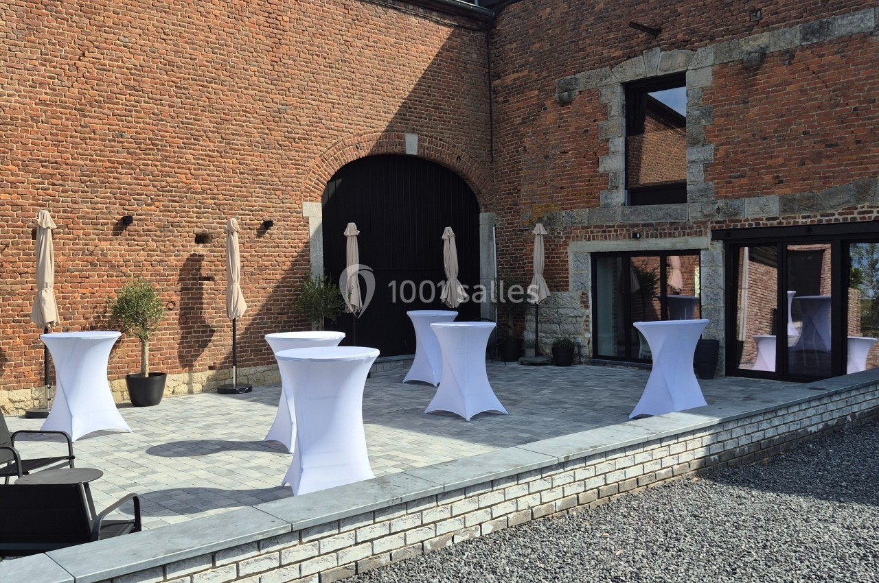 Terrasse en pavés avec tables hautes recouvertes de nappes blanches, parasols fermés et murs en briques rouges.