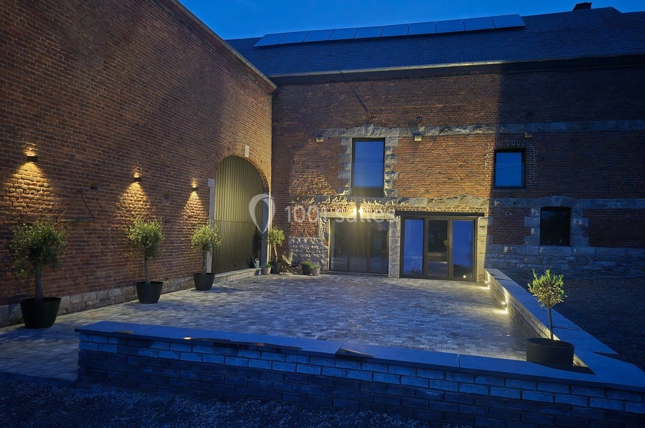 Cour pavée éclairée par des appliques murales, entourée de murs en briques et de pots avec des arbustes.