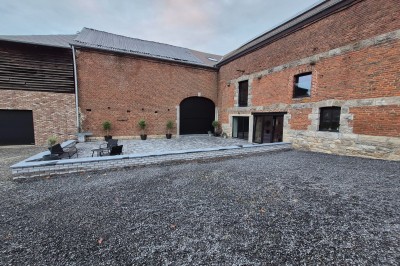 Salle spacieuse avec murs en pierre, poutres en bois apparentes et sol en carrelage gris, éclairée par des fenêtres.