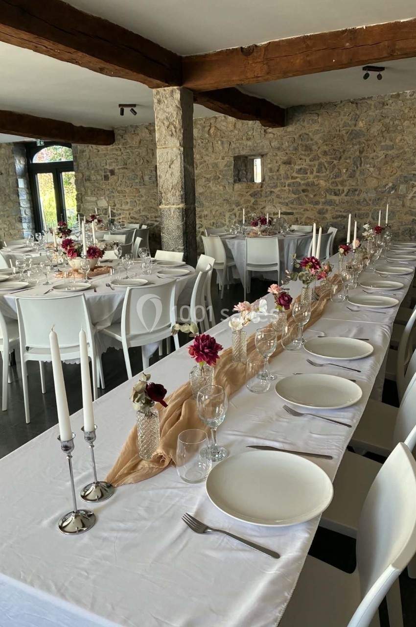 Salle de réception avec tables dressées, nappes blanches, bougies et décorations florales dans un cadre en pierre apparente.