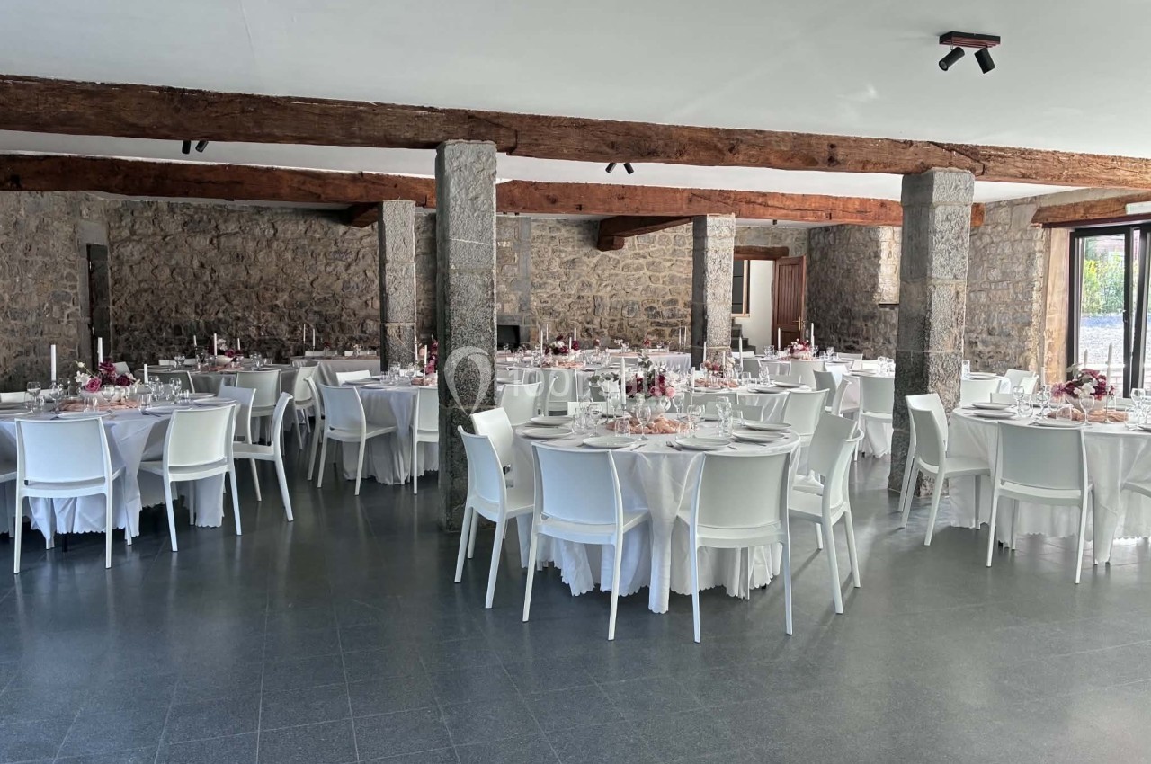 Salle de réception avec des tables rondes dressées, nappes blanches, chaises modernes et murs en pierre apparente.