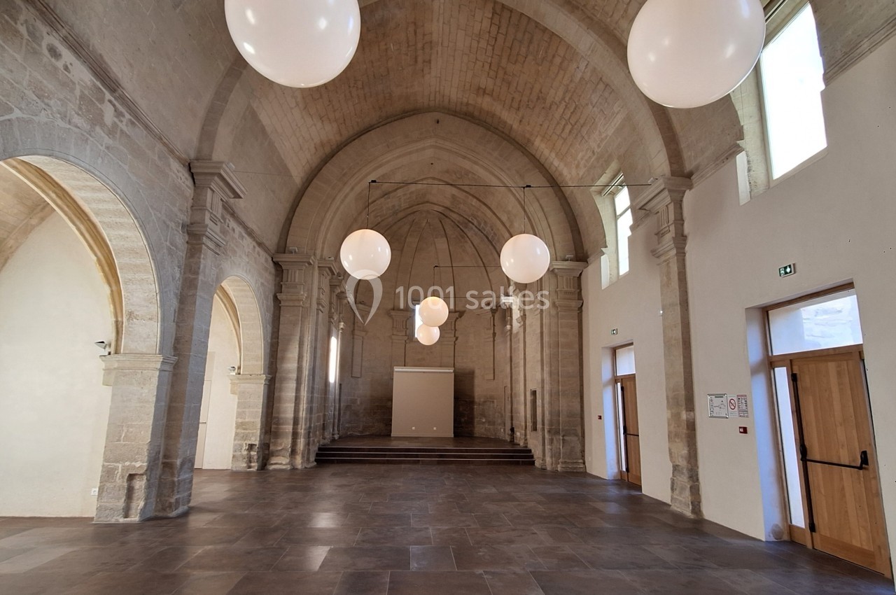 Salle voûtée en pierre avec des arches, des suspensions lumineuses rondes et des portes en bois.