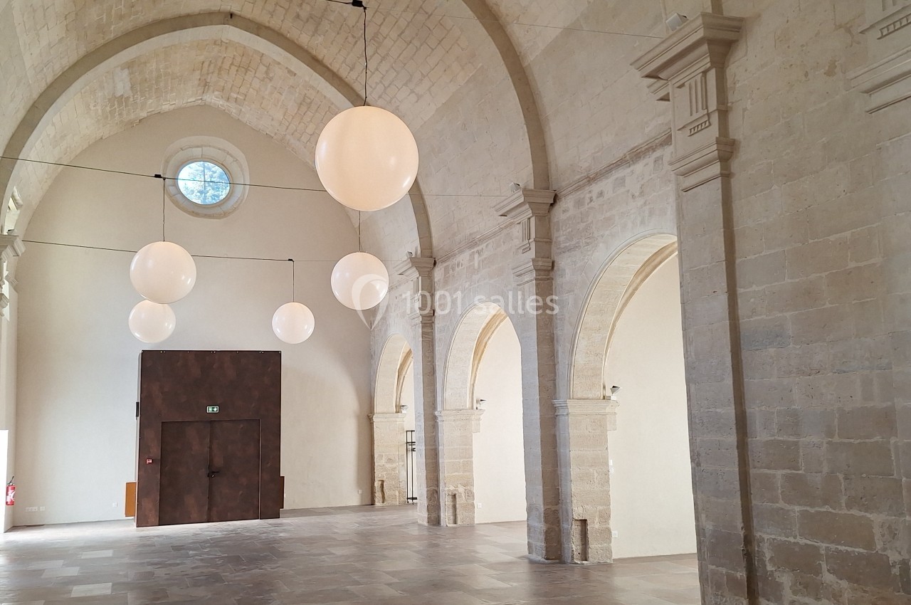 Salle voûtée en pierre avec arches, grandes suspensions lumineuses rondes et porte en bois sombre.