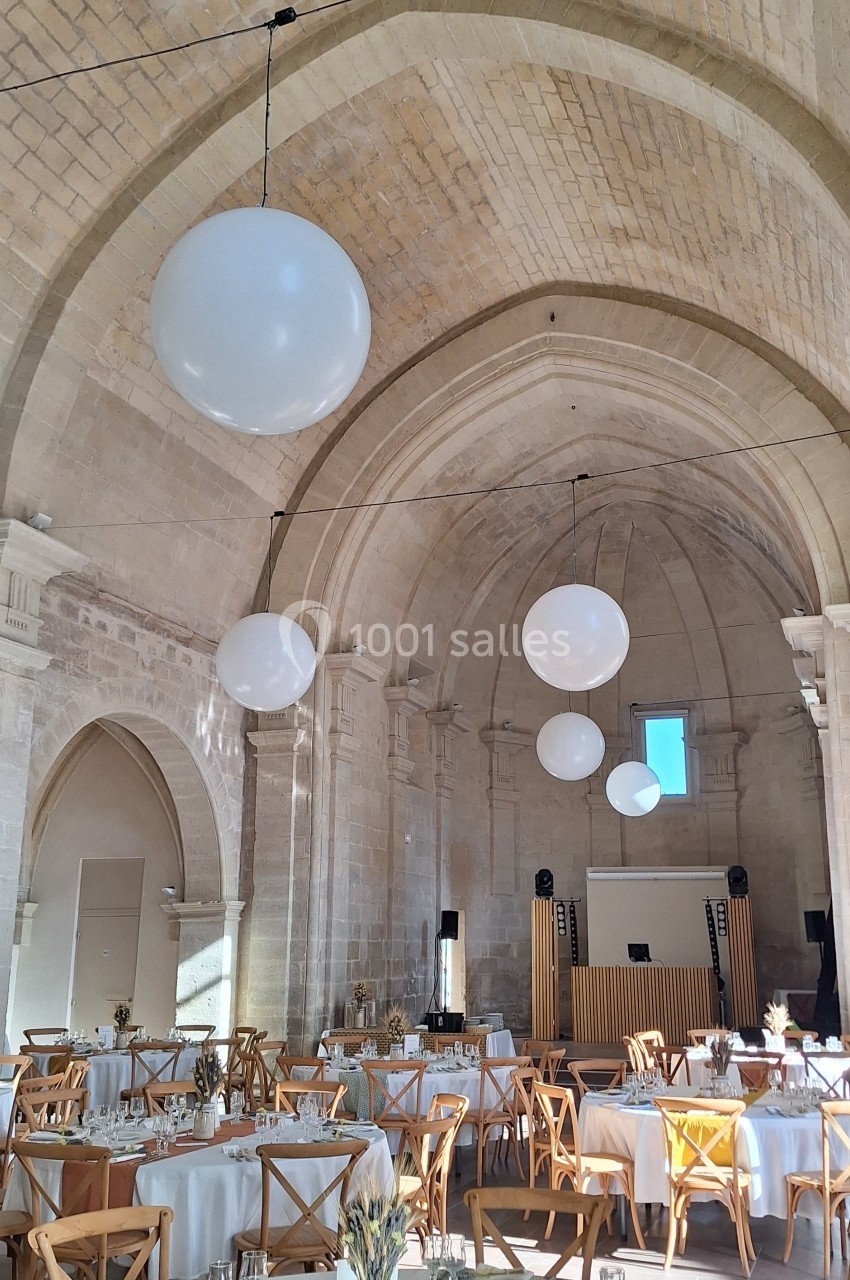 Salle voûtée lumineuse décorée pour un événement, avec tables dressées et grandes suspensions rondes blanches.