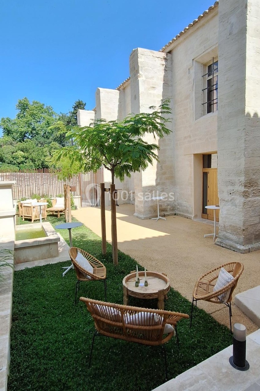 Cour extérieure avec mobilier en rotin, petite fontaine, arbres et façade en pierre sous un ciel bleu.