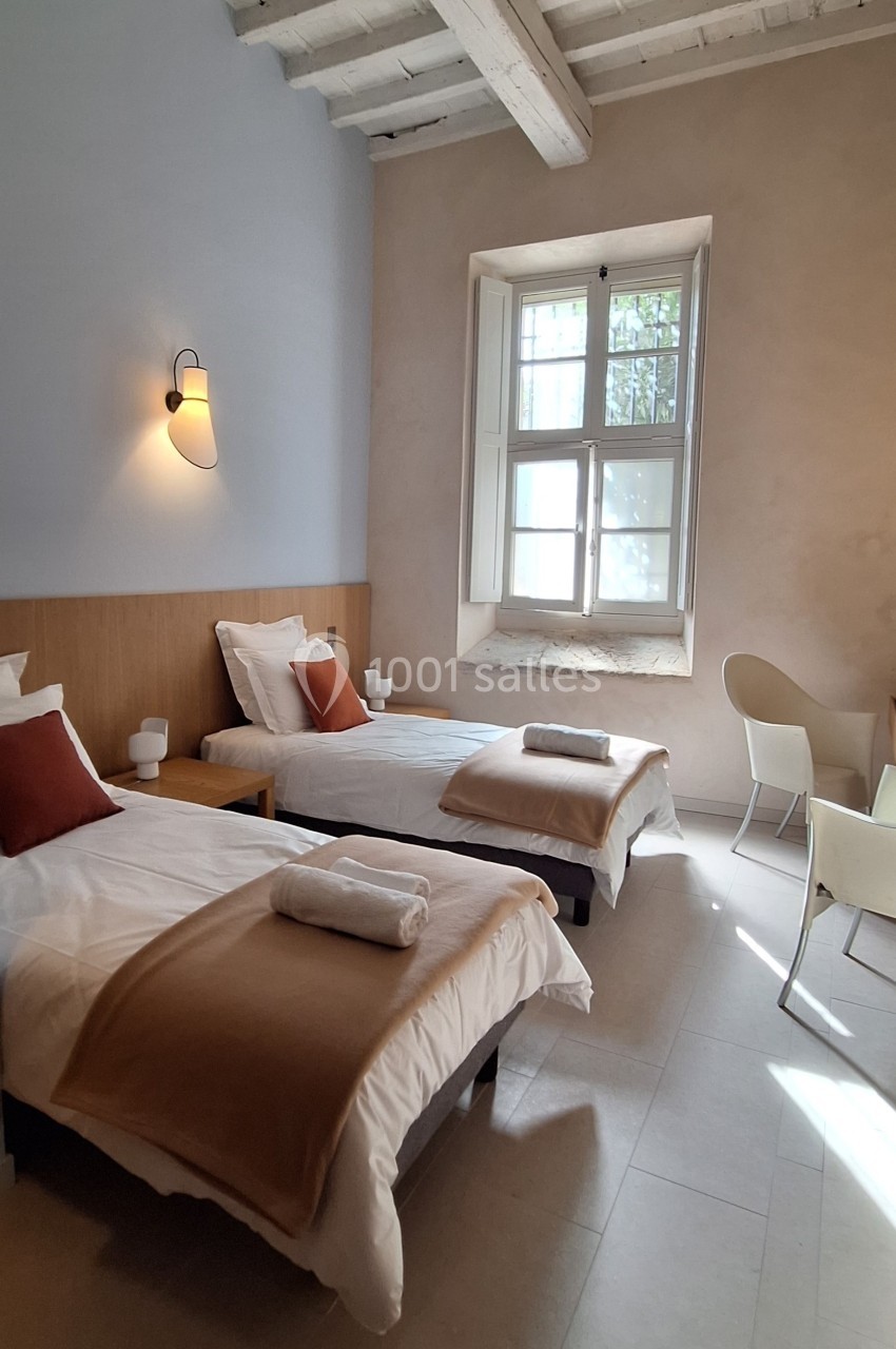 Deux lits simples avec linge beige et oreillers rouges dans une chambre lumineuse avec fenêtre et plafond en bois.