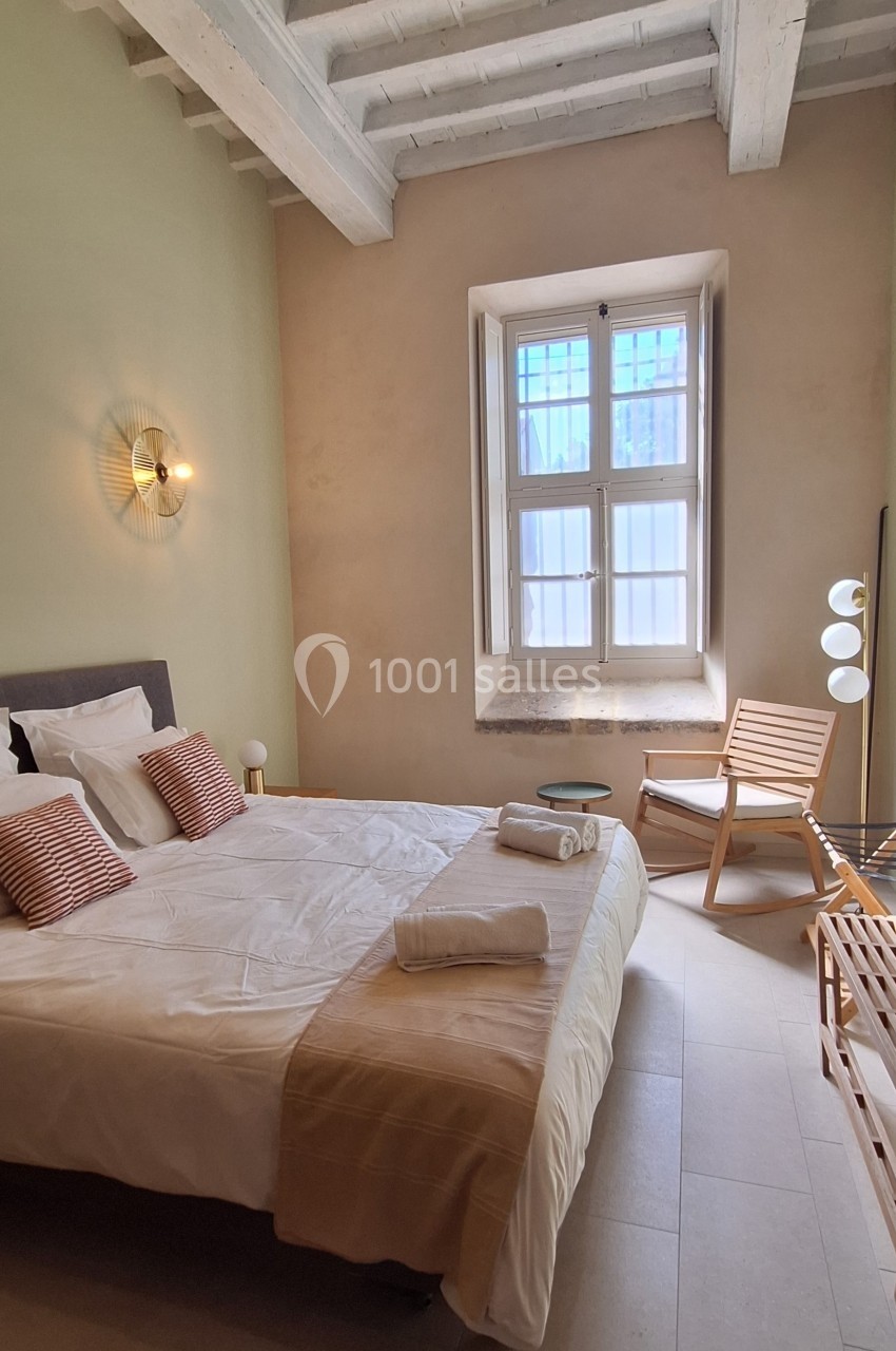 Chambre lumineuse avec lit double, murs clairs, plafond en poutres apparentes, chaise en bois et fenêtre à petits carreaux.