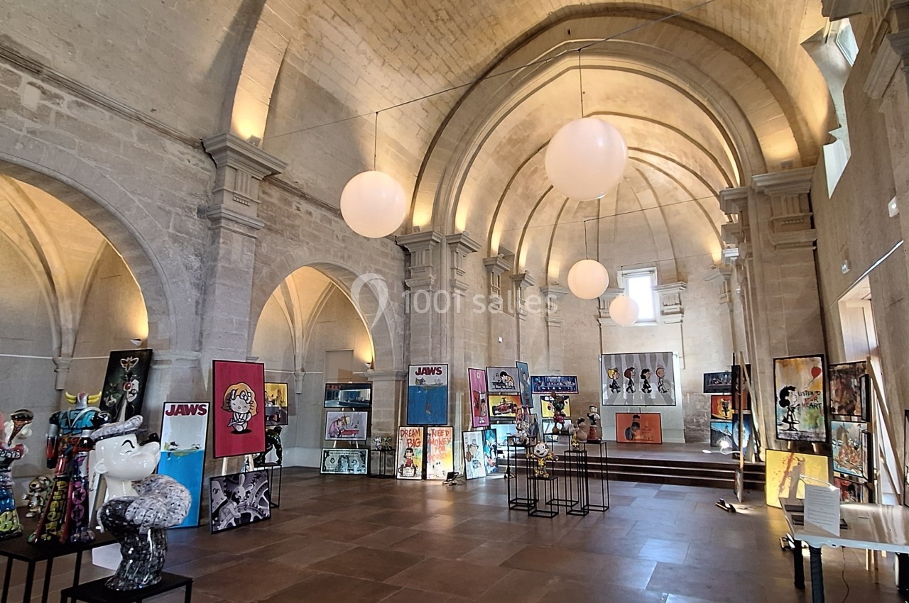 Salle voûtée avec exposition d'art contemporain, sculptures et peintures colorées, éclairage doux et visiteurs.