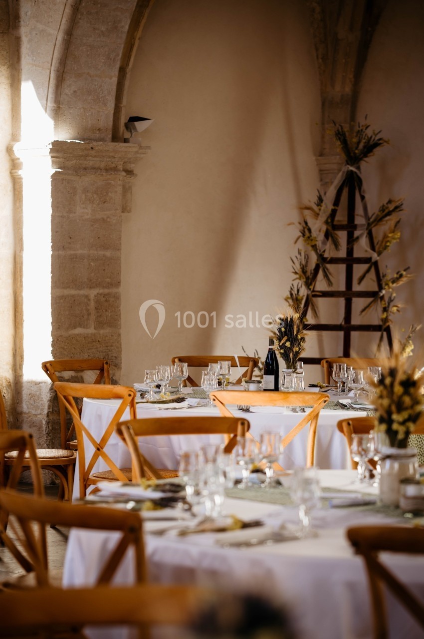 Salle décorée pour un événement avec tables rondes, nappes blanches, chaises en bois et arrangements floraux.
