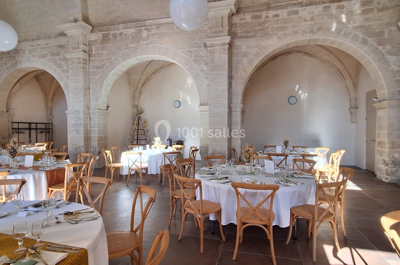 Salle de réception lumineuse avec tables rondes dressées, chaises en bois et décorations élégantes dans un cadre en pierre.