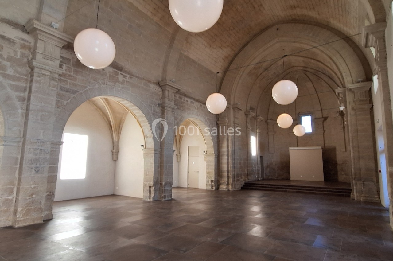 Salle voûtée en pierre avec arches, éclairée par des suspensions rondes et des fenêtres latérales.