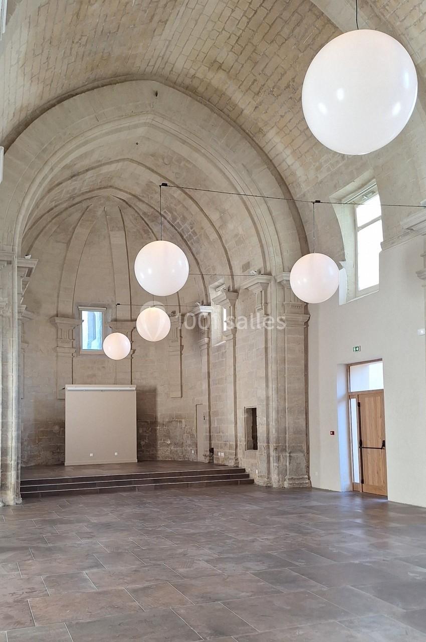 Salle voûtée en pierre avec des suspensions lumineuses rondes et un sol en dalles, éclairée par des fenêtres.