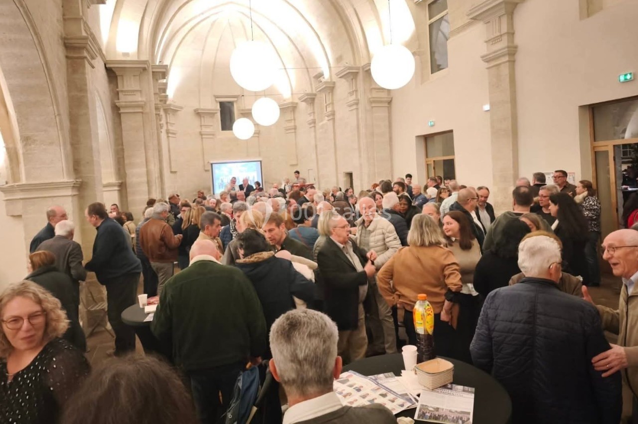 Groupe de personnes réunies dans une grande salle voûtée, discutant autour de tables hautes.