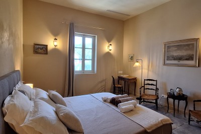 Chambre lumineuse avec lit double, bureau, fauteuils en bois, lampes et fenêtre donnant sur un paysage extérieur.