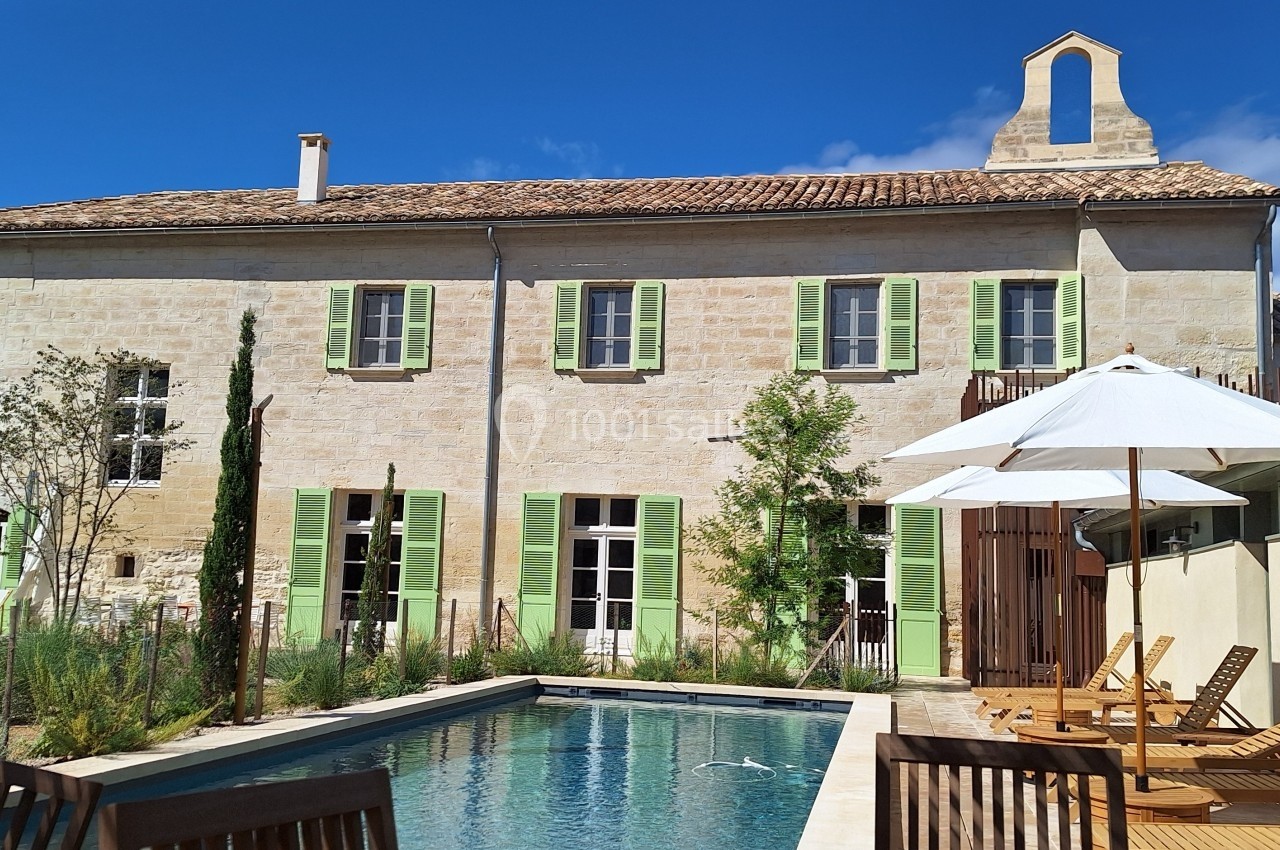 Façade en pierre d'une maison avec volets verts, piscine au premier plan et chaises longues sous parasols.