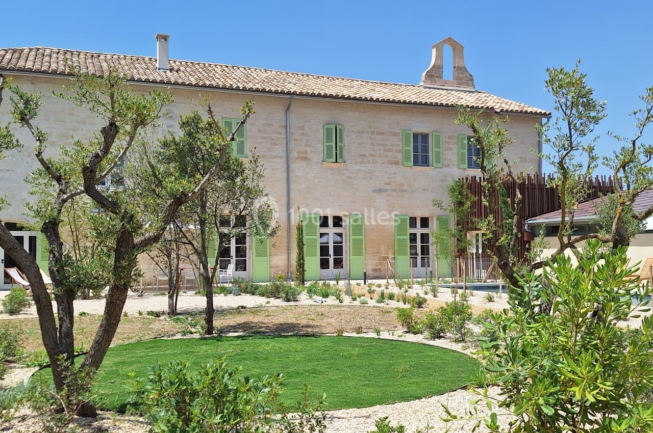 Façade d'une maison en pierre avec volets verts, jardin aménagé et piscine en arrière-plan sous un ciel bleu.