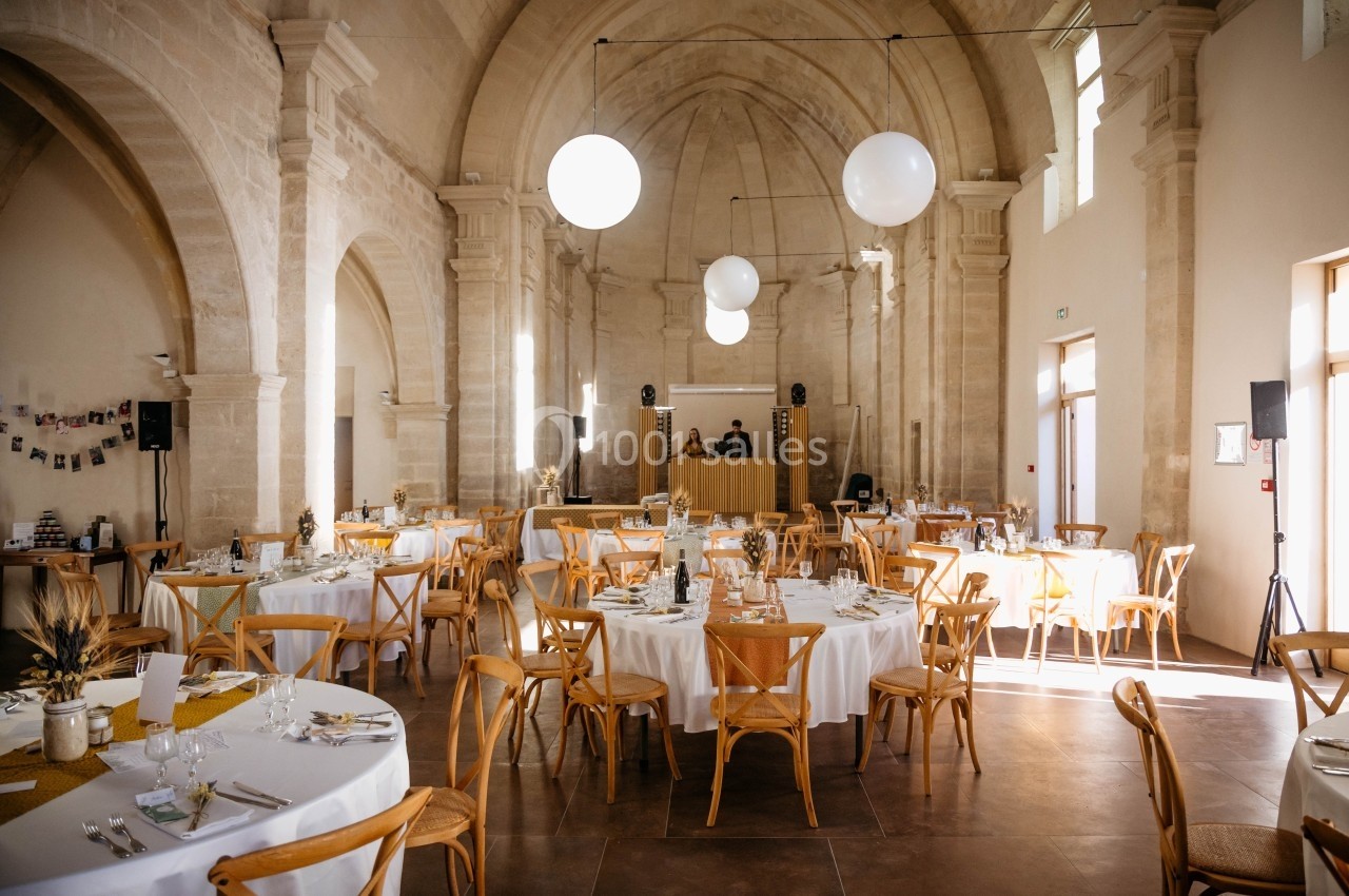 Salle de réception lumineuse avec tables rondes dressées, chaises en bois et décorations élégantes sous une voûte en pierre.