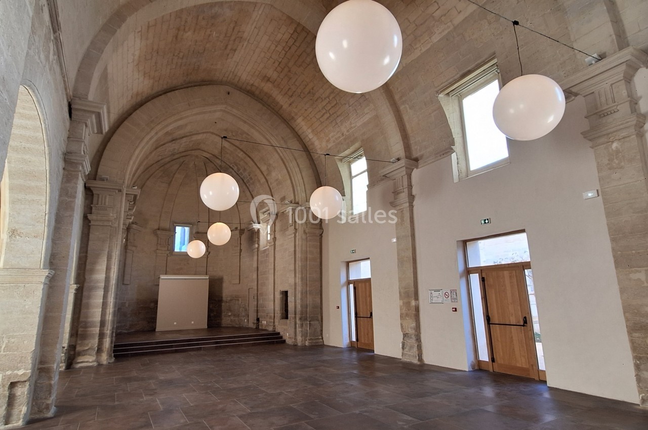 Intérieur d'une salle voûtée en pierre avec des arches, des portes en bois et des luminaires sphériques suspendus.