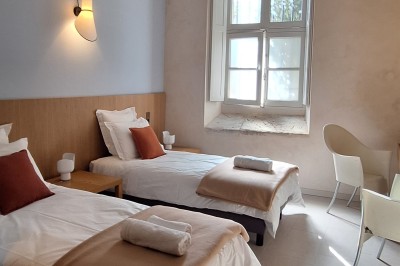 Chambre lumineuse avec lit double, bureau, fauteuils en bois, lampes et fenêtre donnant sur un paysage extérieur.