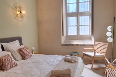 Chambre lumineuse avec lit double, bureau, fauteuils en bois, lampes et fenêtre donnant sur un paysage extérieur.