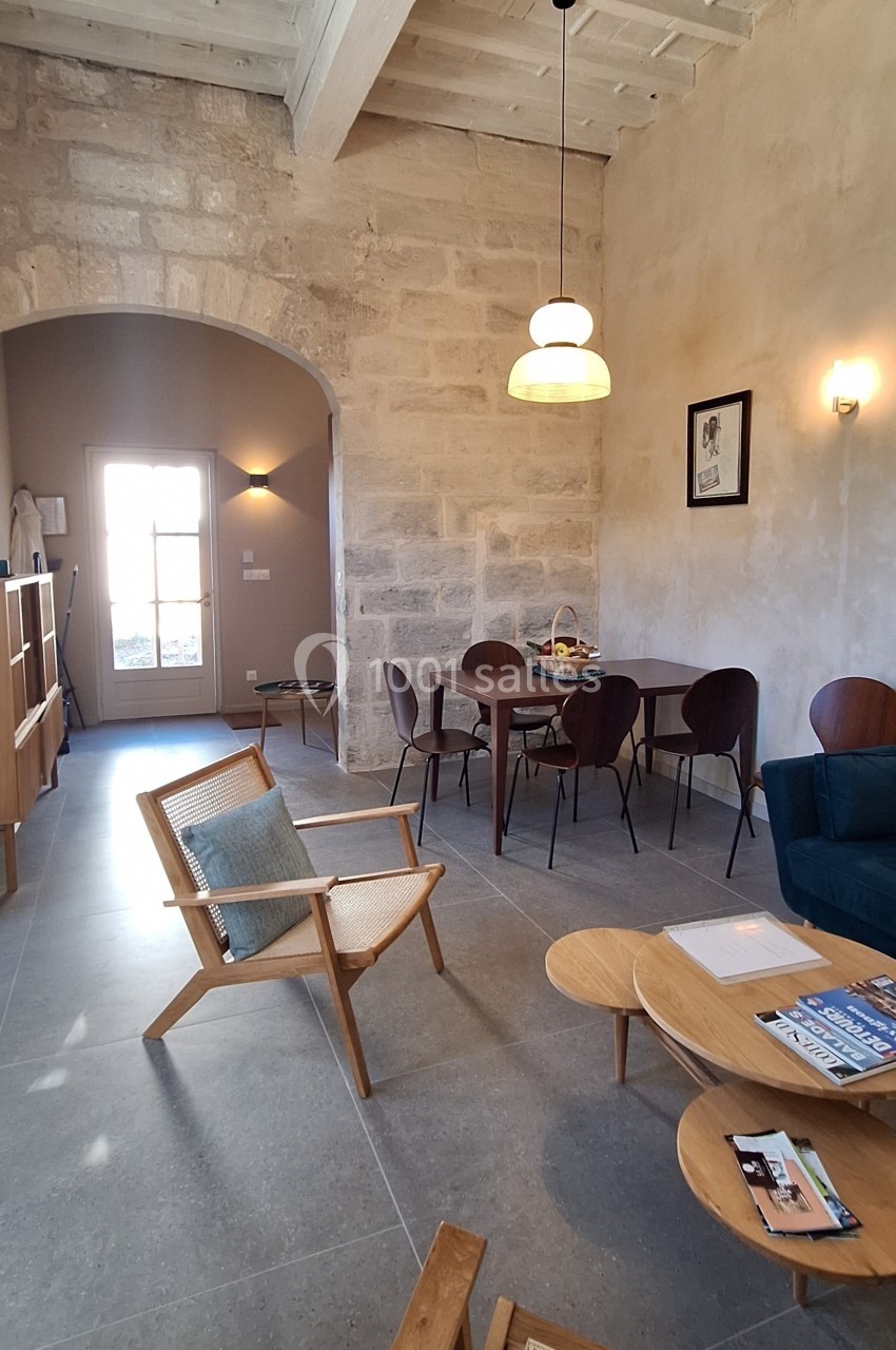 Salle de séjour lumineuse avec murs en pierre, mobilier en bois, table à manger, fauteuils et éclairage suspendu.