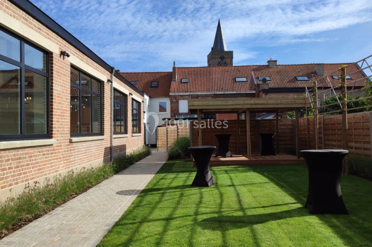 Jardin aménagé avec pelouse, terrasse en pavés, tables hautes et pergola, entouré de bâtiments en briques.