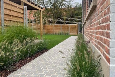 Jardin aménagé avec pelouse, terrasse en pavés, tables hautes et pergola, entouré de bâtiments en briques.