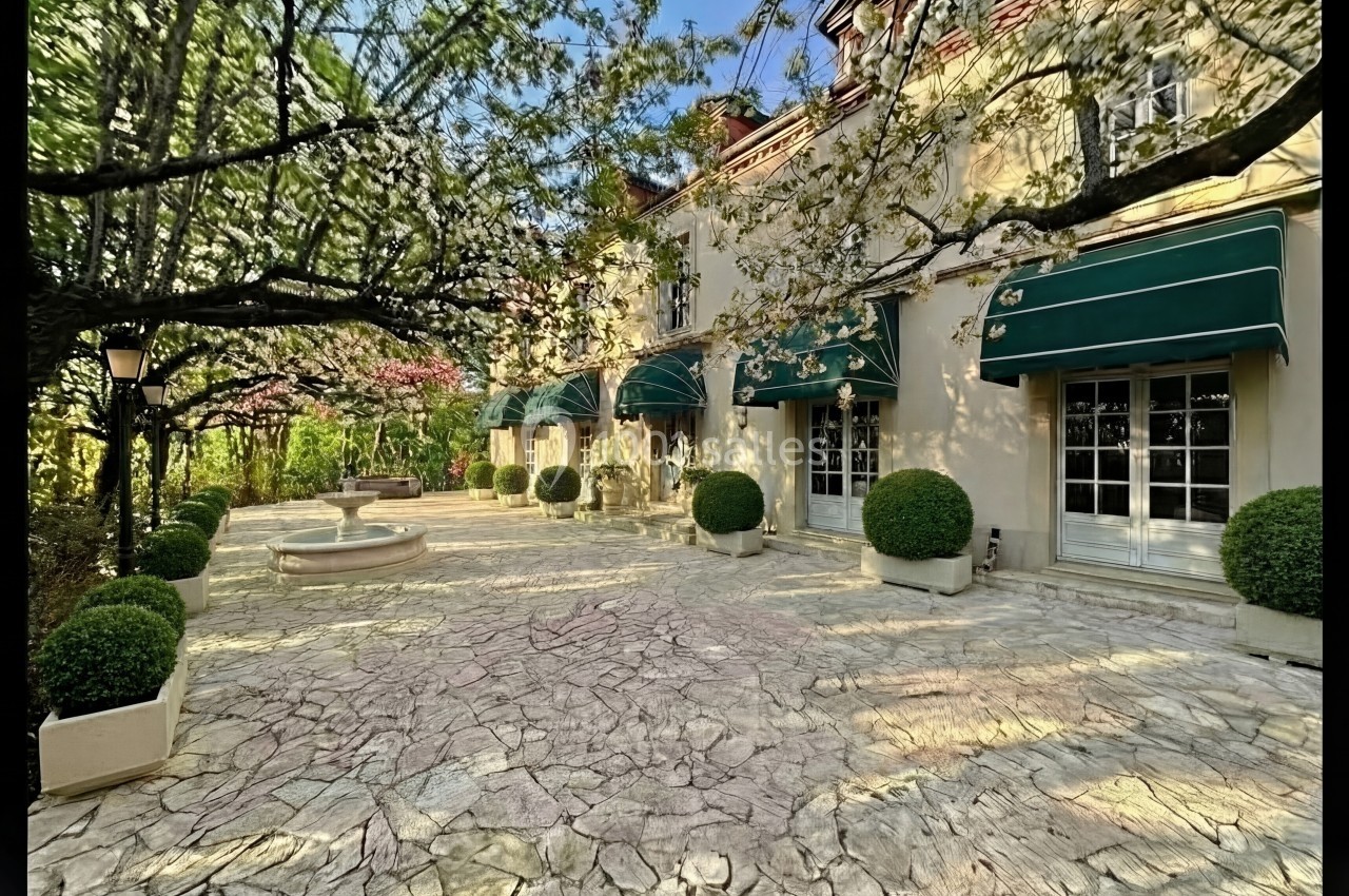 Cour pavée bordée d'arbres en fleurs, avec une fontaine centrale et une façade ornée de stores verts.