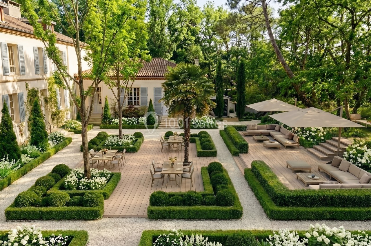 Jardin aménagé avec terrasses en bois, mobilier d'extérieur, haies taillées, fleurs blanches et arbres environnants.