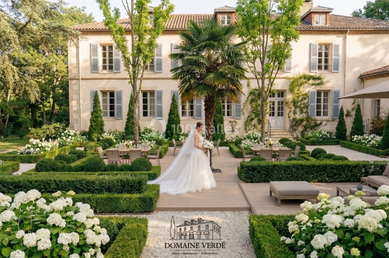 Une mariée en robe blanche marche dans un jardin élégant devant une grande maison de style classique.