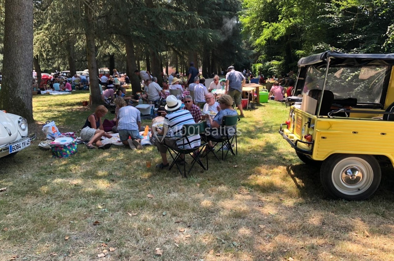 Des personnes pique-niquent en groupe dans un parc boisé, avec des tables, des chaises et une voiture jaune à proximité.