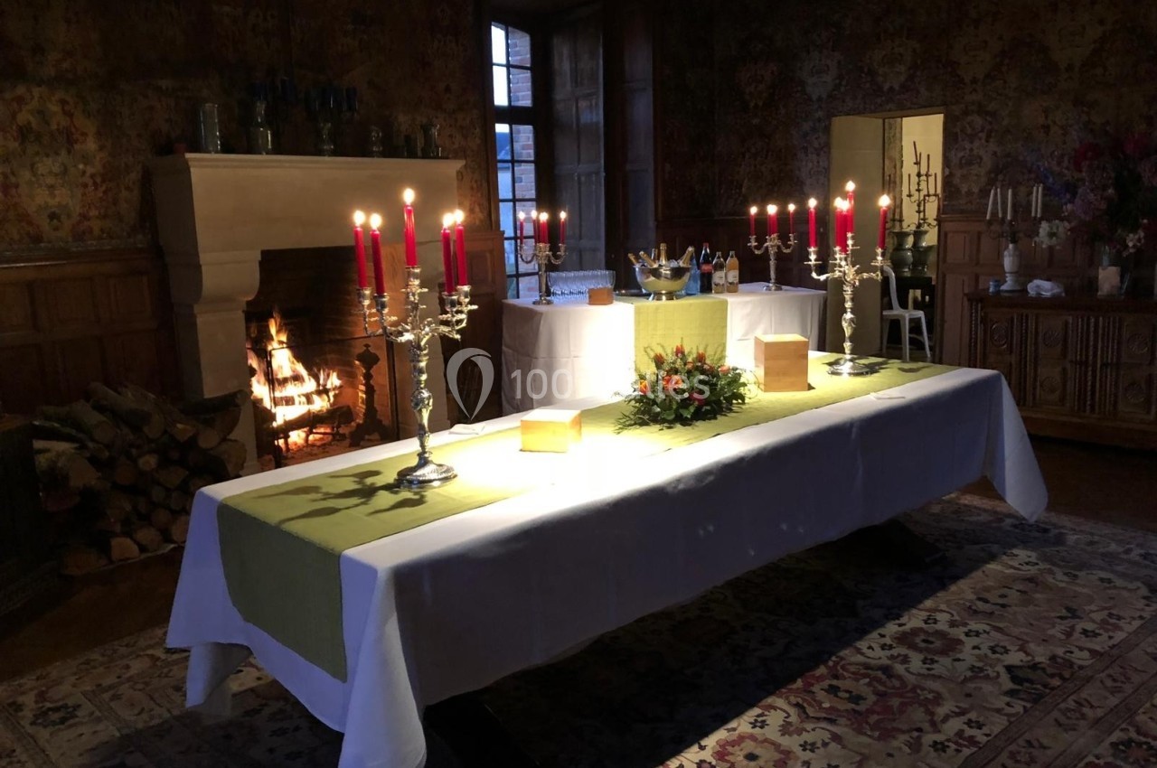 Table dressée avec nappes blanches, chandeliers allumés et décoration florale, dans une pièce avec cheminée et tapis.