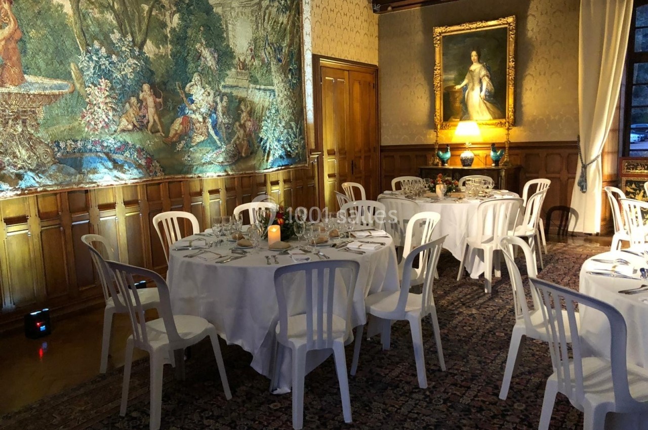 Salle élégante avec tables rondes dressées, chaises blanches, tapis, tapisserie murale et tableau éclairé.