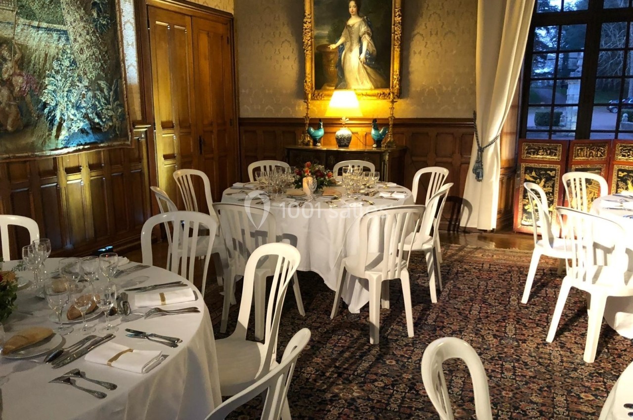 Salle élégante avec tables rondes dressées, chaises blanches, tapis décoratif, tableau et lumière tamisée.