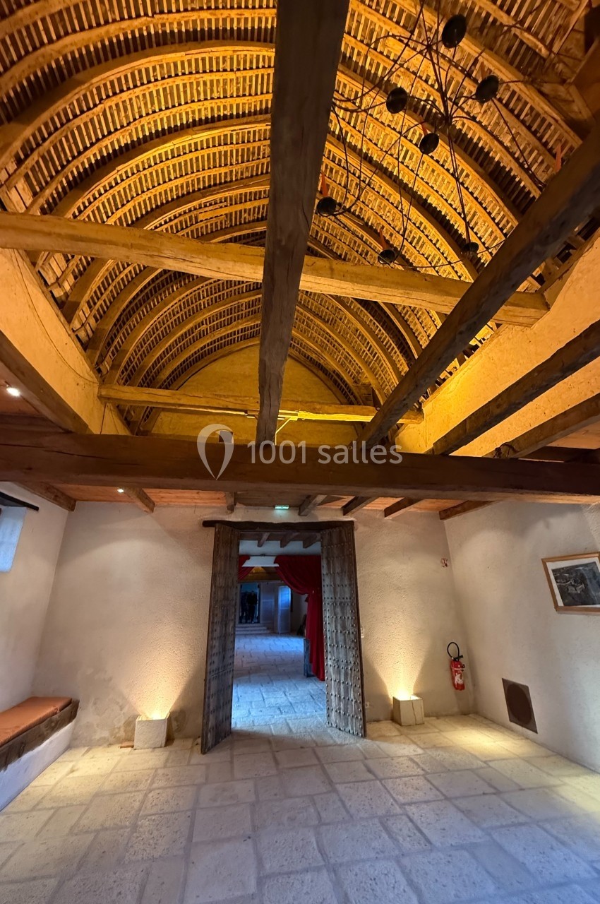 Plafond voûté en bois avec poutres apparentes dans une salle en pierre éclairée, vue vers une porte ouverte.