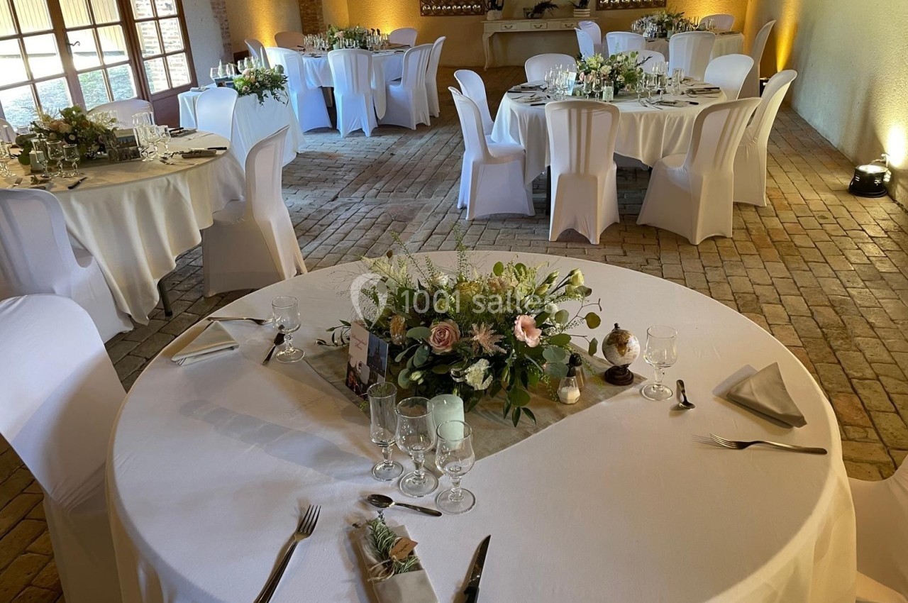 Salle de réception avec tables rondes décorées de nappes blanches, fleurs et vaisselle élégante sur un sol en briques.