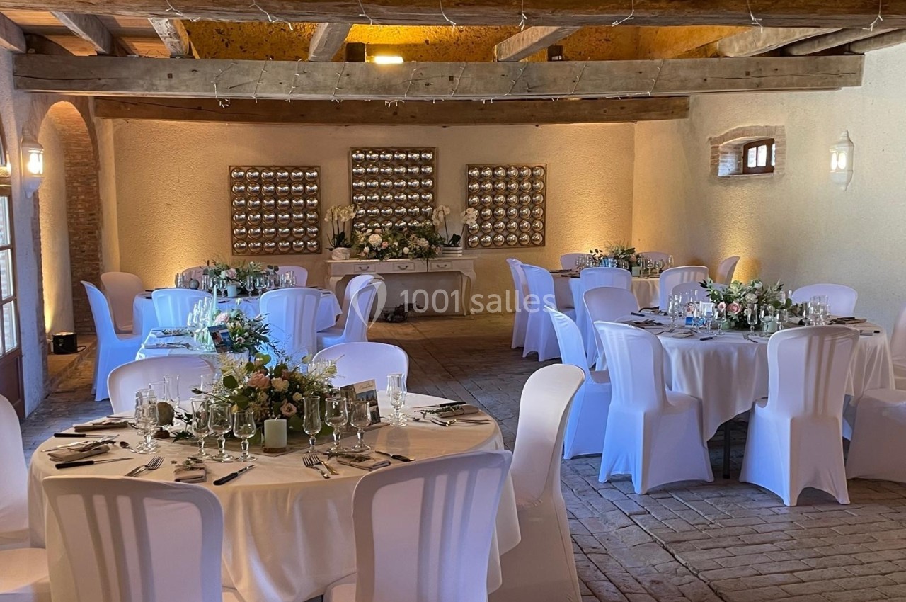 Salle de réception décorée avec des tables rondes dressées, nappes blanches et arrangements floraux, éclairage chaleureux.