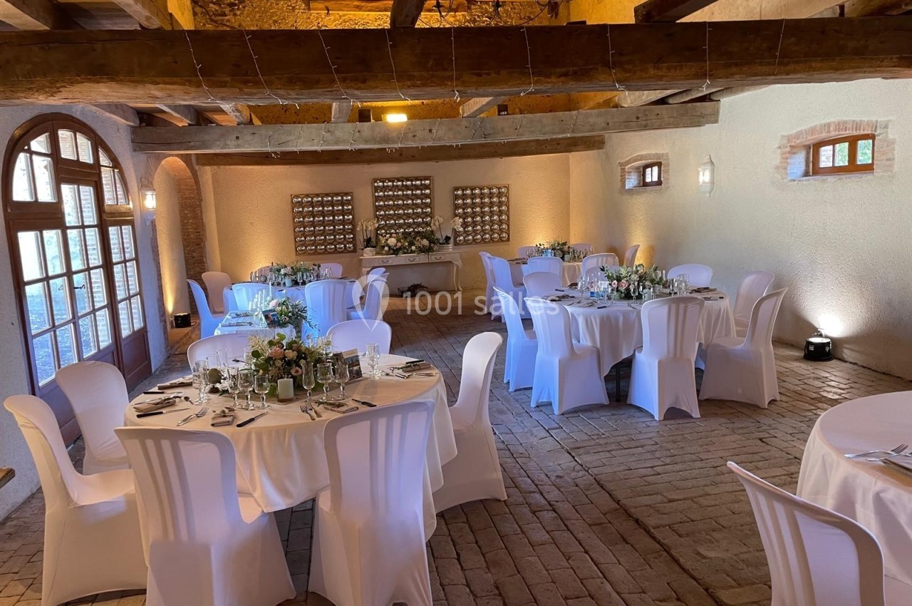 Salle de réception avec tables rondes décorées, chaises blanches et poutres apparentes dans un cadre rustique.