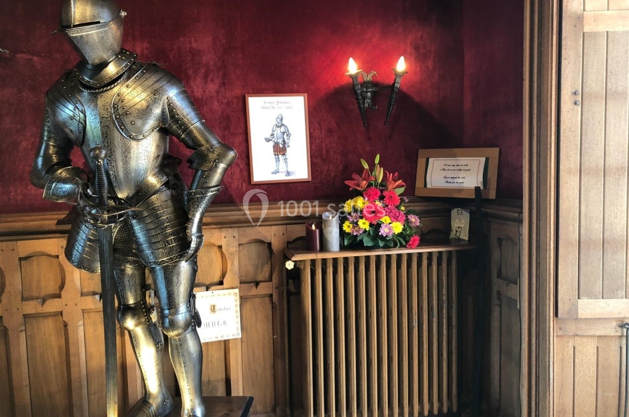 Armure médiévale exposée dans une pièce aux murs rouges, avec un bouquet de fleurs et des cadres accrochés.