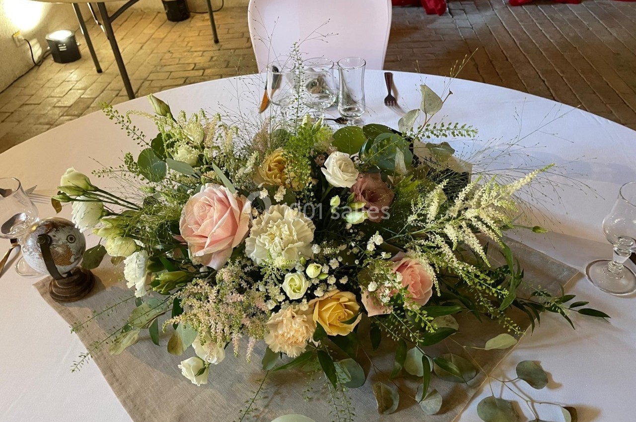 Centre de table floral avec roses, feuillages et fleurs variées sur une nappe beige, entouré de couverts et verres.