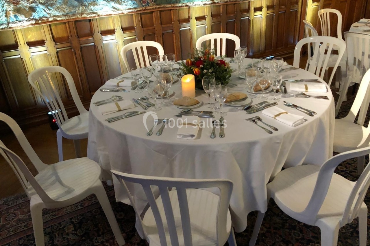 Table ronde dressée avec une nappe blanche, couverts, verres, bougie et centre de table floral, entourée de chaises blanches.