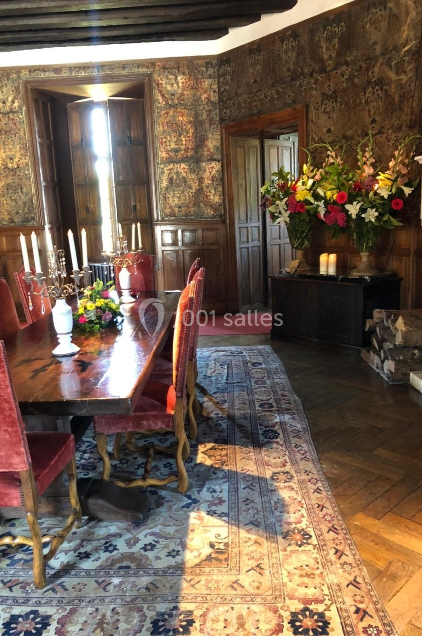 Salle à manger avec table en bois, chaises rouges, tapis persan, cheminée et bouquet de fleurs colorées.