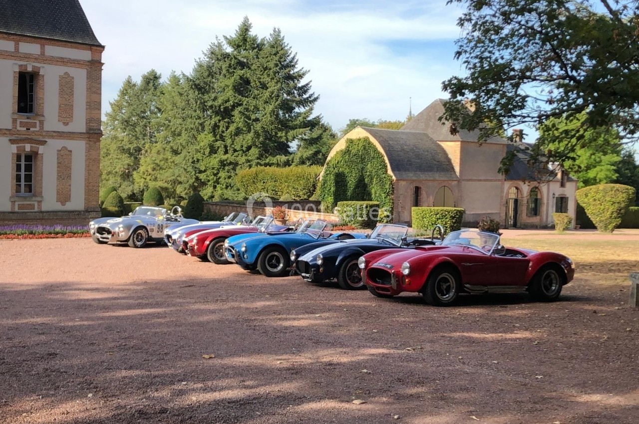 Alignement de voitures anciennes colorées stationnées devant un bâtiment historique entouré de verdure.