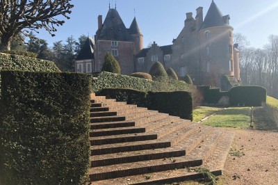 Château en briques rouges entouré de pelouses et d'arbres, sous un ciel clair et ensoleillé.